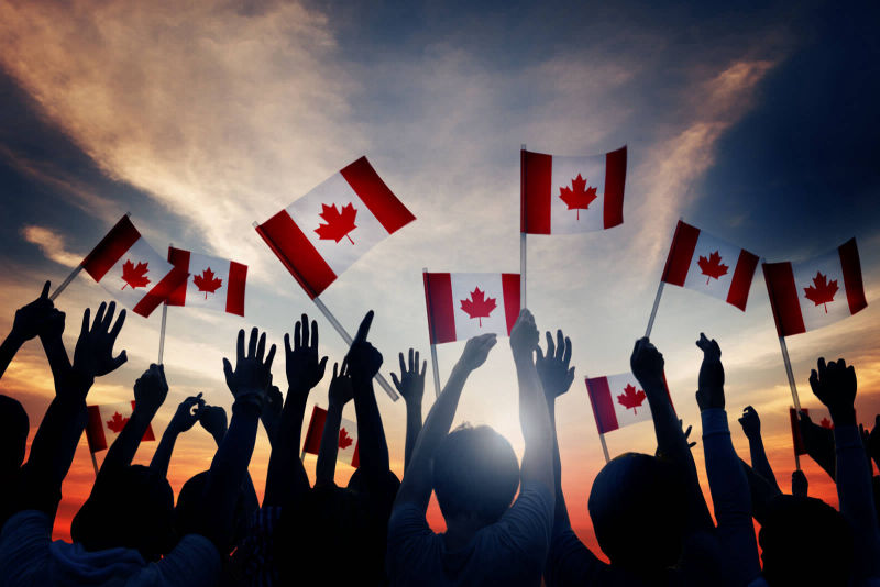A crowd of people waving the Canadian flag and throwing their hands in the air in fun and freedom.  The backdrop is beautiful pink and orange sunset with streaked clouds.