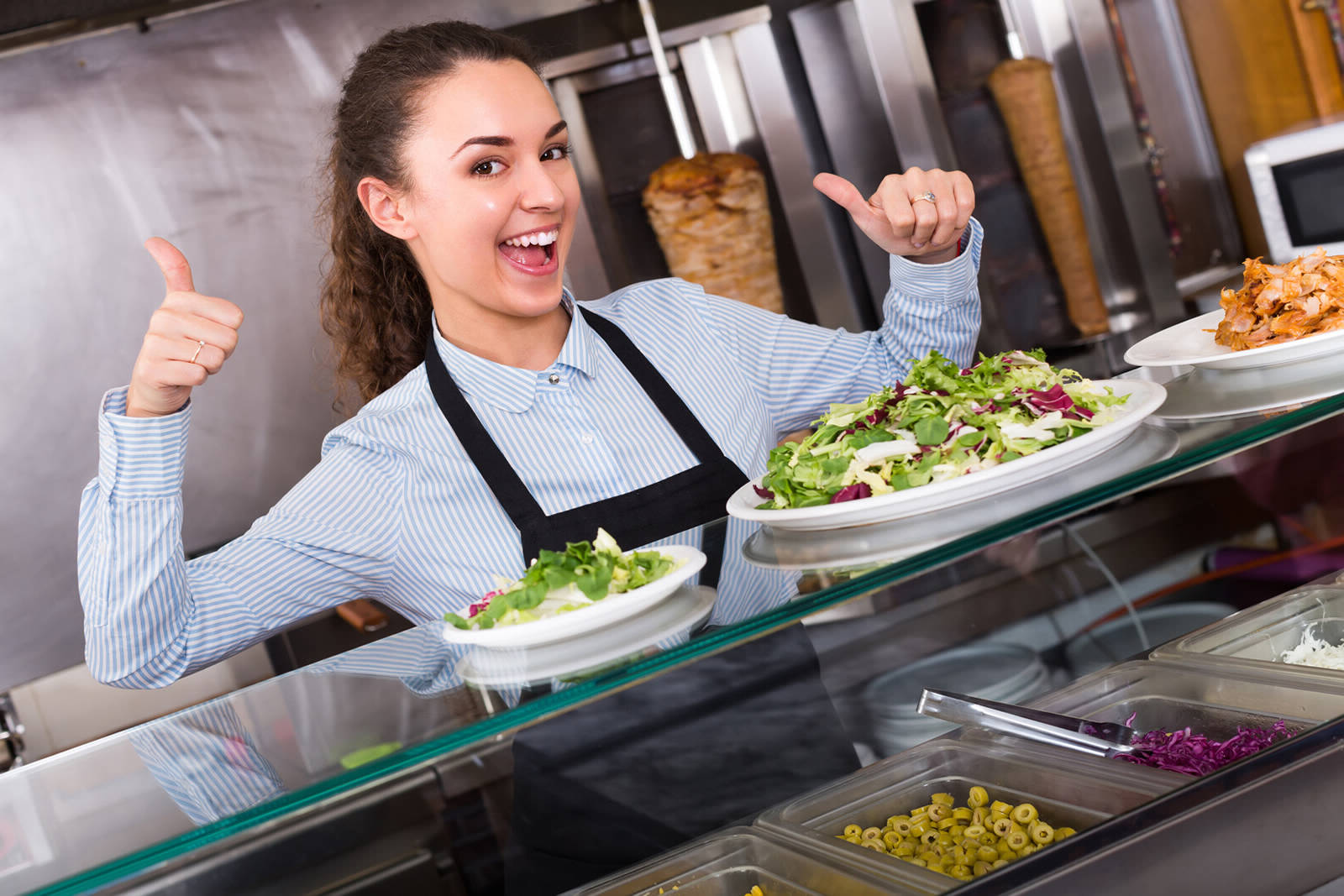 A restaurant server with her thumbs up behind the service line in the kitchen, with a Caesar Salad and another fresh salad on the counter.