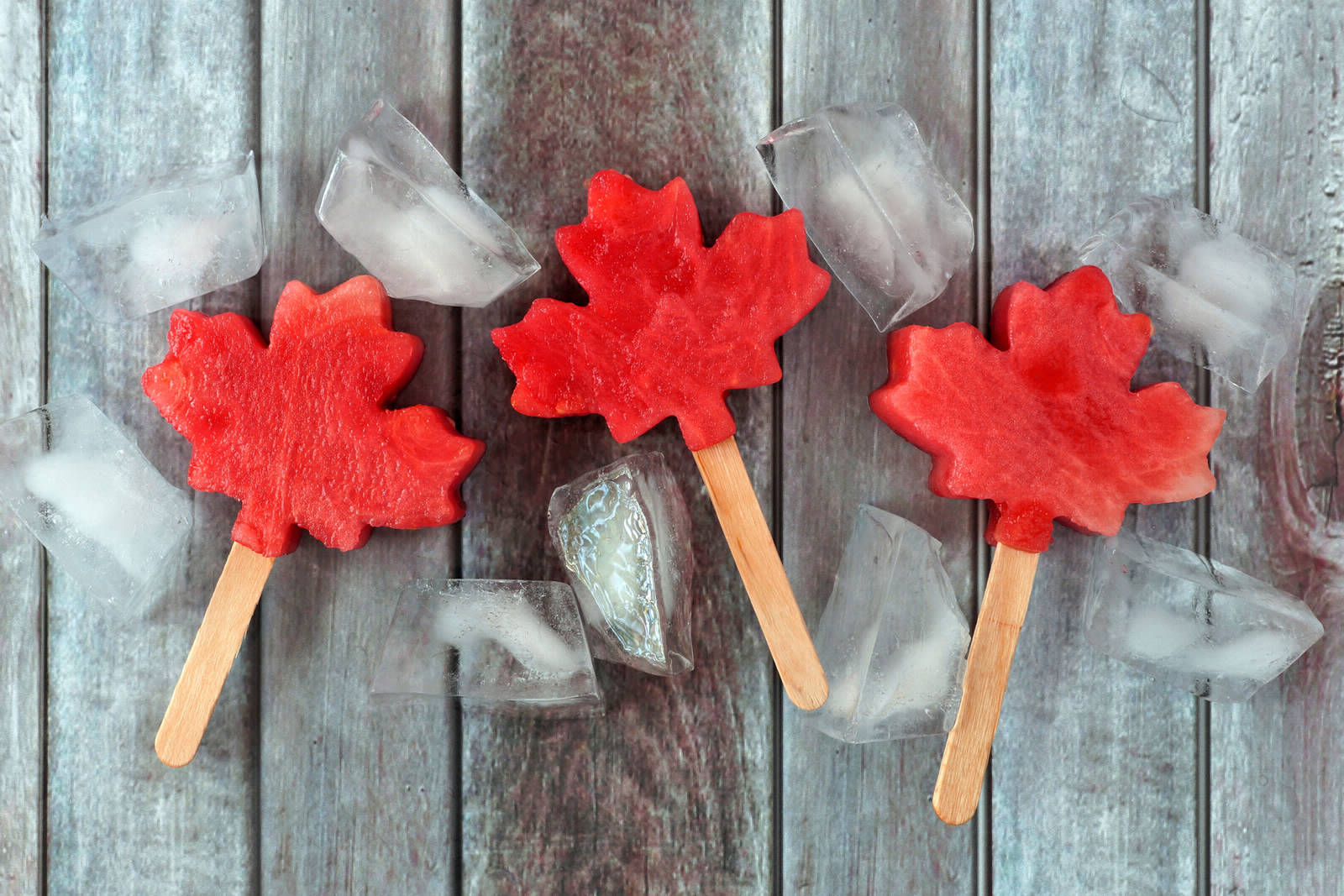 Three red maple leaf shaped popsicles on a grey barn board background with several ice cubes around them.